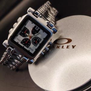 May include: A silver Oakley watch with a square face and a stainless steel band. The watch face has black sub-dials with red hands and the Oakley logo. The watch is resting on a silver container with the Oakley logo.