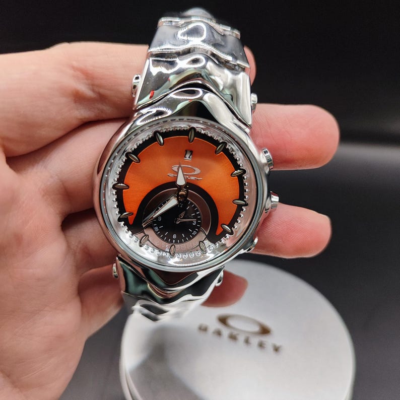 Pu&ograve; includere: Un orologio da polso Oakley argentato con quadrante arancione e nero. L'orologio ha un cinturino unico e strutturato e un quadrante rotondo con pi&ugrave; quadranti. Il logo Oakley &egrave; visibile sul quadrante e sulla cassa dell'orologio.