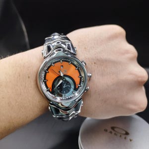 Pu&ograve; includere: Un orologio da polso argentato con quadrante arancione e nero. L'orologio ha un design scolpito unico ed &egrave; indossato al polso. Il quadrante presenta il logo del marchio. Un accessorio alla moda.