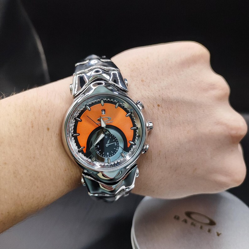 Pu&ograve; includere: Un orologio da polso argentato con quadrante arancione e nero. L'orologio ha un design scolpito unico ed &egrave; indossato al polso. Il quadrante presenta il logo del marchio. Un accessorio alla moda.