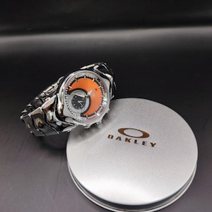Pu&ograve; includere: Un orologio Oakley argentato con quadrante arancione e nero e cinturino argentato abbinato. L'orologio &egrave; accanto a una scatola di latta argentata con il marchio Oakley.