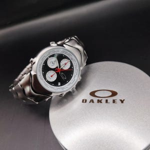May include: A silver-toned Oakley watch with a black face, white subdials, and red accents. The watch is displayed next to a silver Oakley box with the brand's logo.
