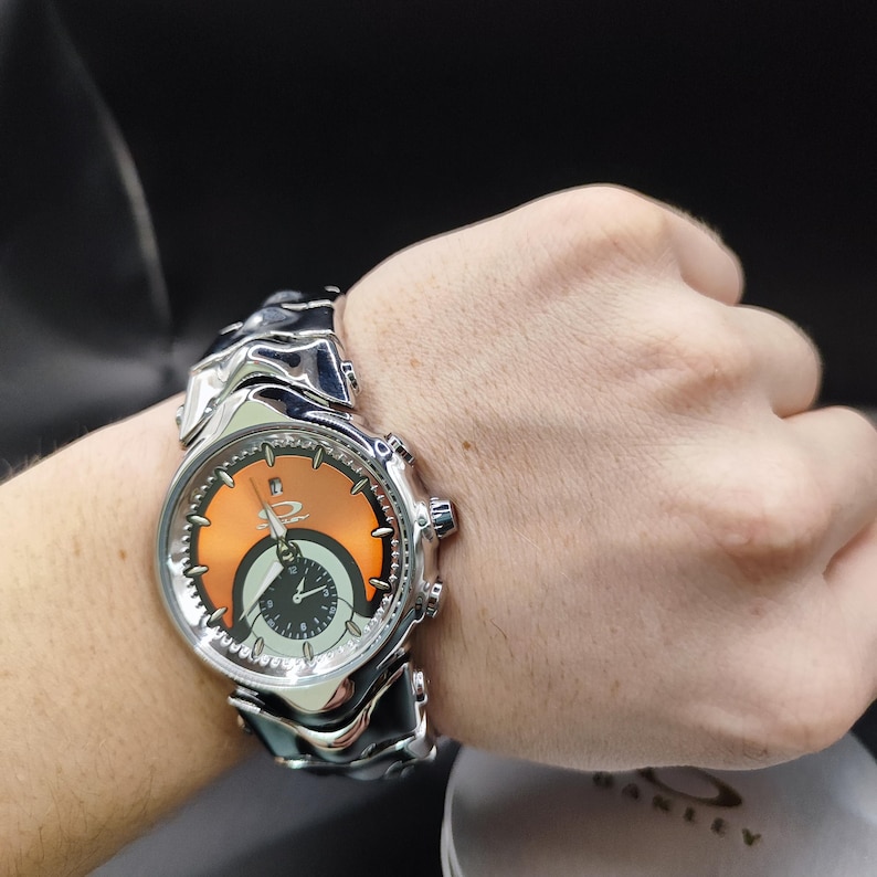 Pu&ograve; includere: Un orologio da polso argentato con quadrante arancione e nero &egrave; mostrato su un polso. L'orologio ha un cinturino unico e strutturato e diversi quadranti. Il quadrante dell'orologio presenta il logo del marchio. L'orologio &egrave; un accessorio elegante.