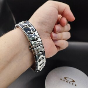 Pu&ograve; includere: Un bracciale argentato con un design segmentato unico. Il bracciale &egrave; composto da maglie rettangolari con un motivo strutturato e ondulato. La chiusura &egrave; visibile e il bracciale &egrave; mostrato su un polso. Il logo Oakley &egrave; visibile sullo sfondo.