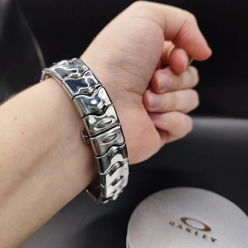Pu&ograve; includere: Un bracciale argentato con un design segmentato unico. Il bracciale &egrave; composto da maglie rettangolari con un motivo strutturato e ondulato. La chiusura &egrave; visibile e il bracciale &egrave; mostrato su un polso. Il logo Oakley &egrave; visibile sullo sfondo.