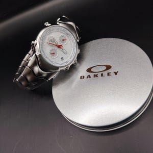 Puede incluir: Un reloj Oakley plateado con esfera blanca y detalles rojos, junto a una caja plateada con la marca Oakley. El reloj tiene una correa de eslabones y tres subesferas. La caja es redonda y tiene el logotipo de Oakley.