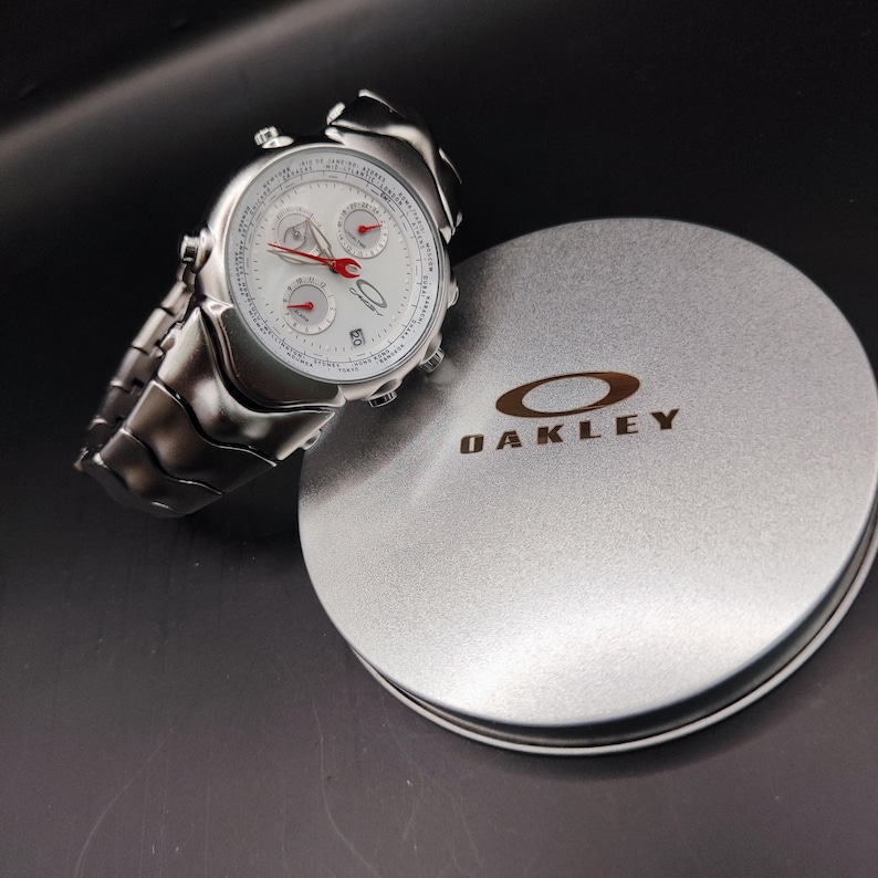 Puede incluir: Un reloj Oakley plateado con esfera blanca y detalles rojos, junto a una caja plateada con la marca Oakley. El reloj tiene una correa de eslabones y tres subesferas. La caja es redonda y tiene el logotipo de Oakley.