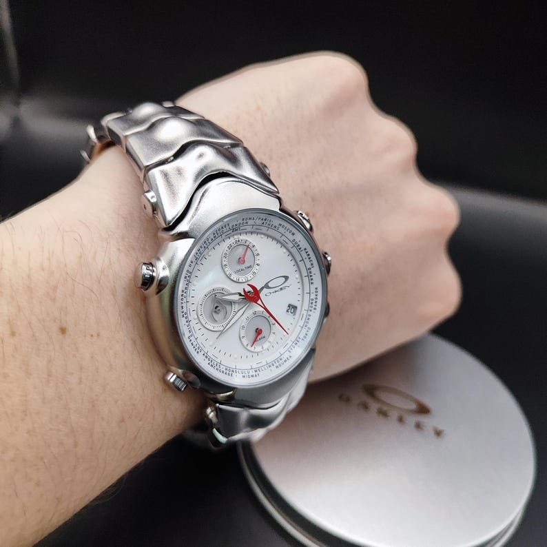 Puede incluir: Un reloj plateado con esfera blanca y detalles rojos se muestra en una mu&ntilde;eca. El reloj presenta m&uacute;ltiples subesferas y una pantalla de fecha. La correa del reloj es de metal plateado a juego. El reloj est&aacute; junto a un recipiente Oakley plateado.