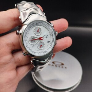 Puede incluir: Un reloj Oakley plateado con esfera blanca, manecillas rojas y m&uacute;ltiples subesferas. El reloj tiene una correa texturizada y se muestra con su estuche de marca. La esfera muestra la fecha y las zonas horarias.