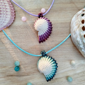 May include: Two macrame shell pendants with cord necklaces. One pendant is white and purple, the other is white and teal. The necklaces are on a wooden surface with seashells and beads.