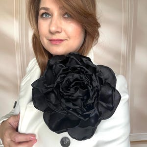 May include: A large, black satin flower brooch pinned to a white coat. The flower is the focal point, with multiple layers of petals. The coat has a button closure. The overall aesthetic is elegant and sophisticated.