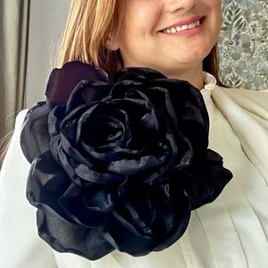 Może przedstawiać: Duża, czarna, wielowarstwowa broszka z tkaniny w kształcie kwiatu, przypięta do kremowej bluzki z wysokim kołnierzem. Kwiat składa się z licznych nakładających się na siebie, półprzezroczystych i nieprzezroczystych płatków o zaokrąglonym kształcie.