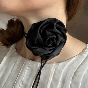 May include: A black satin rose choker necklace. The large, three-dimensional flower is the focal point, with delicate petals and a detailed center. The choker is secured with thin black cords.