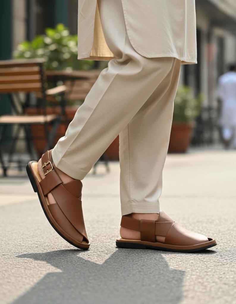 May include: Brown leather sandals with a strap and buckle closure. The sandals have a flat sole and are worn with beige pants. The sandals are designed with a crisscross strap design over the foot.