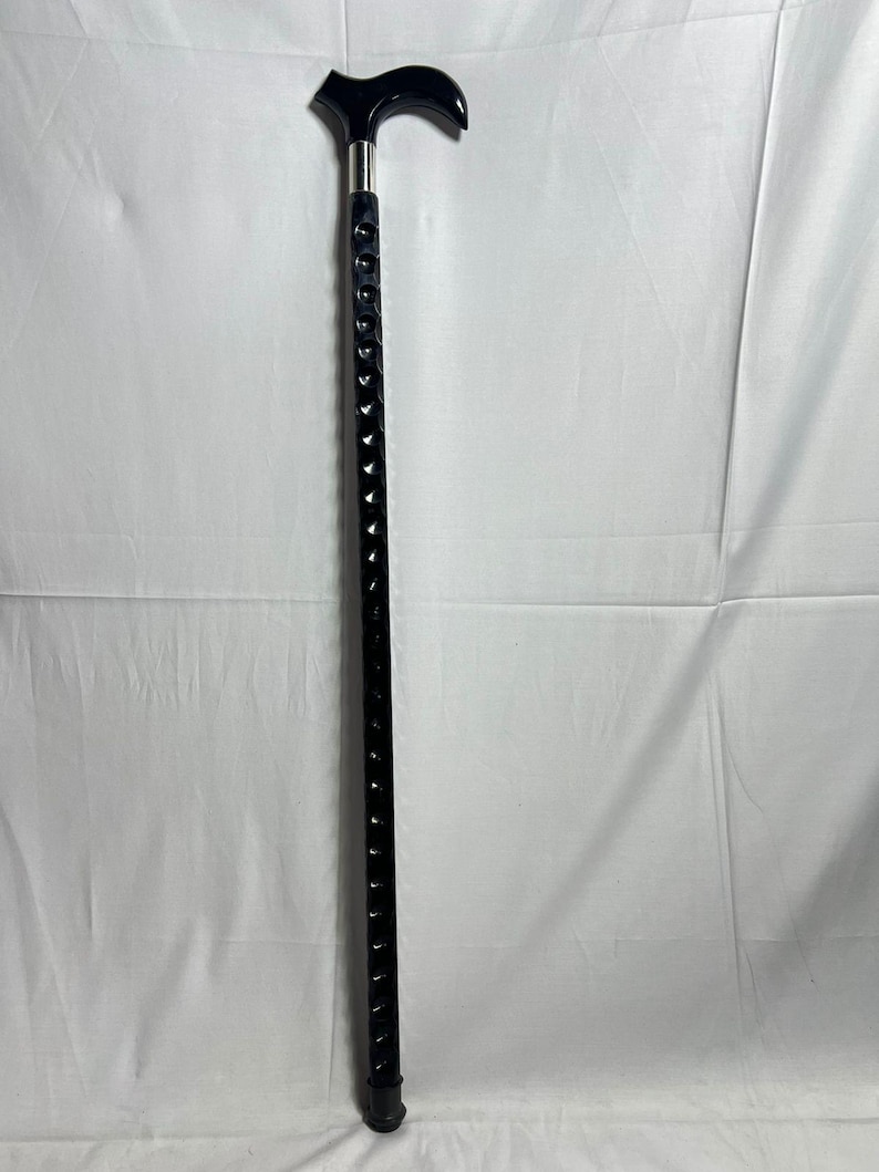 May include: A black walking cane with a curved handle and a silver metal collar. The shaft is black with a series of decorative cutouts. The cane is designed for support and mobility.