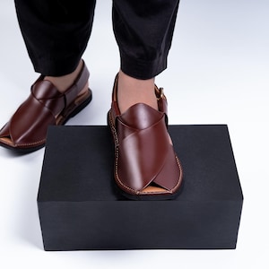 May include: A pair of brown leather sandals with a unique design. The sandals have a strap around the ankle and are displayed on a black box. The leather appears to be stitched with a contrasting thread.