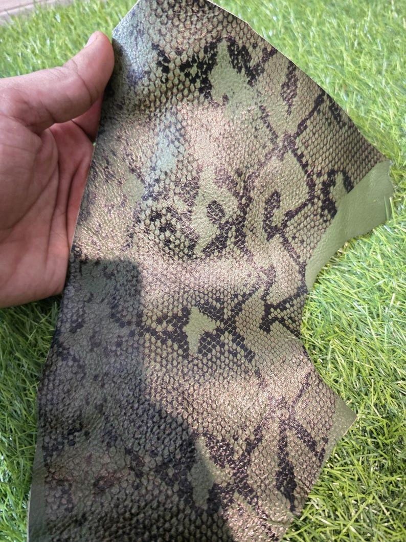 Peut inclure: Un morceau de cuir vert olive avec un motif peau de serpent. Le cuir a une surface textur&eacute;e avec des marques marron fonc&eacute; et noires. Le mat&eacute;riau est pr&eacute;sent&eacute; sur un fond d'herbe verte.