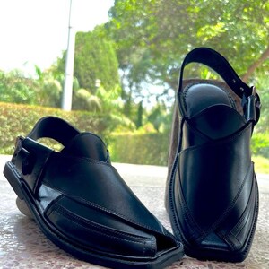 May include: Two black leather sandals with a square toe and a strap that buckles around the ankle. The sandals have a traditional design with stitched details and are set against a blurred green background.