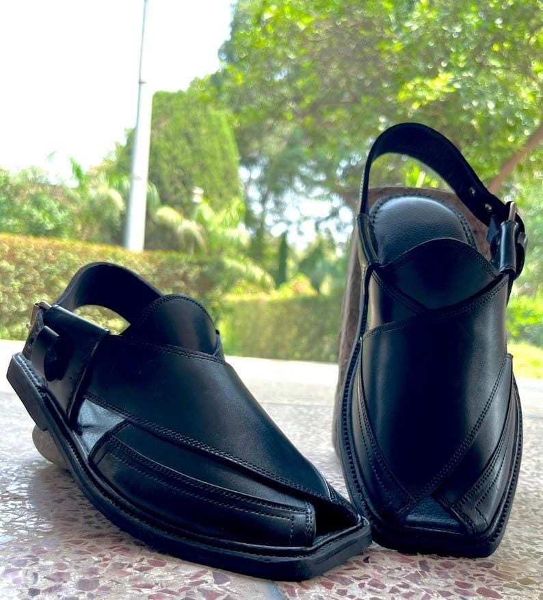 May include: Two black leather sandals with a square toe and a strap that buckles around the ankle. The sandals have a traditional design with stitched details and are set against a blurred green background.
