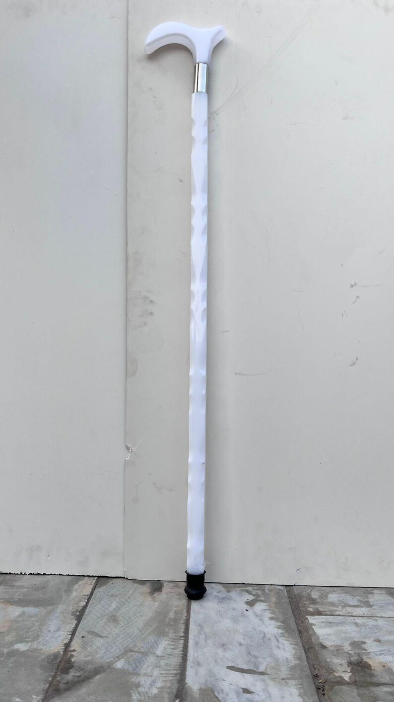 May include: A white walking cane with a curved handle and a textured shaft. The cane has a silver metal band near the handle and a black rubber tip. The cane is approximately 36 inches in length.