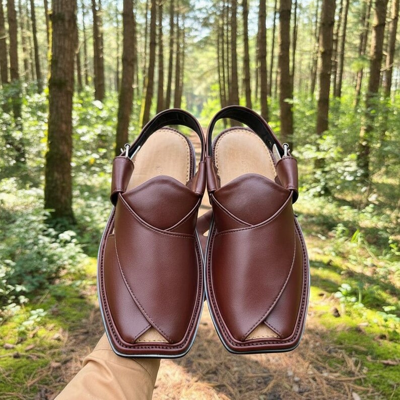 May include: A pair of brown leather Peshawari sandals. These open-toe sandals feature a square toe design, a strap around the ankle, and a leather upper. The sandals are held up in front of a forest background.