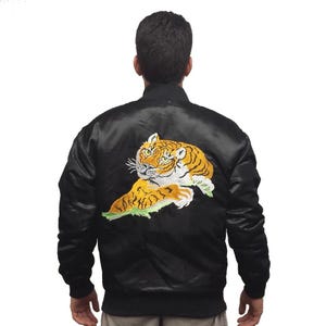 Könnte beinhalten: Schwarze Satin-Bomberjacke mit einem detaillierten, gestickten Tigerdesign auf dem Rücken. Der Tiger hat orange, gelbe und weiße Farben. Die Jacke hat einen klassischen Bomber-Stil mit geripptem Kragen und Bündchen.