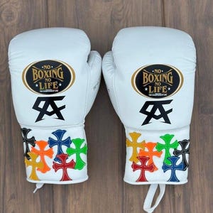 Può includere: Un paio di guantoni da boxe bianchi con una toppa ovale dorata e nera con il testo NO BOXING NO LIFE. Sotto la toppa c'è un logo nero. La zona del polso è decorata con diverse applicazioni colorate a forma di croce in rosso, blu, verde, arancione, giallo e nero.