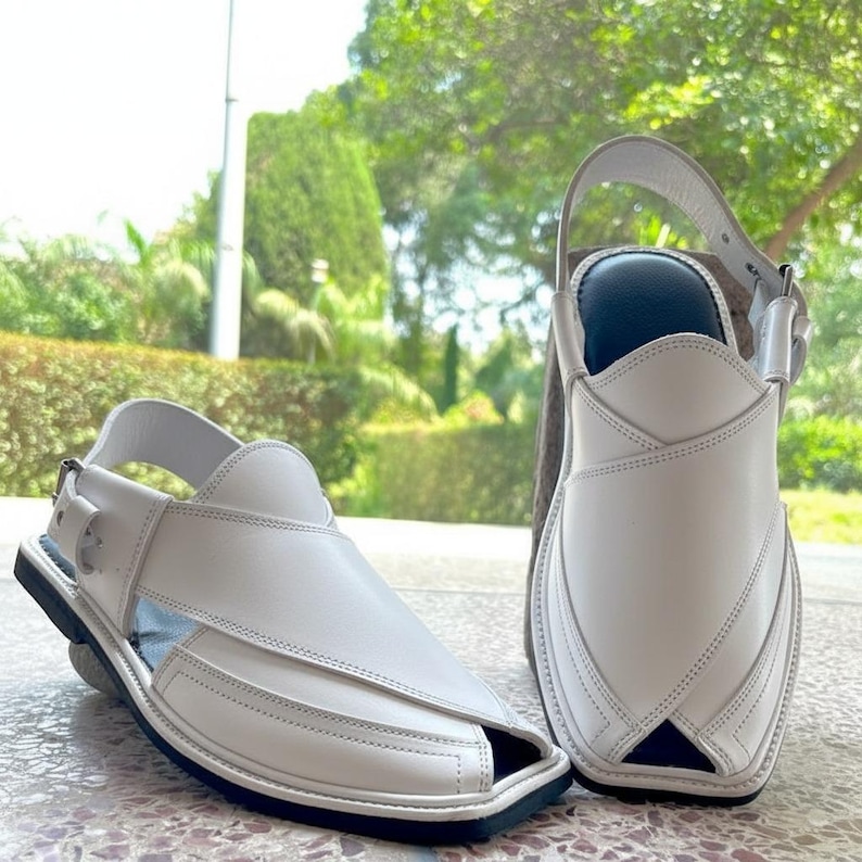 May include: A pair of white leather sandals with a square toe design. The sandals have a strap that goes around the ankle and a buckle closure. The soles are black, and the sandals are sitting on a stone surface.