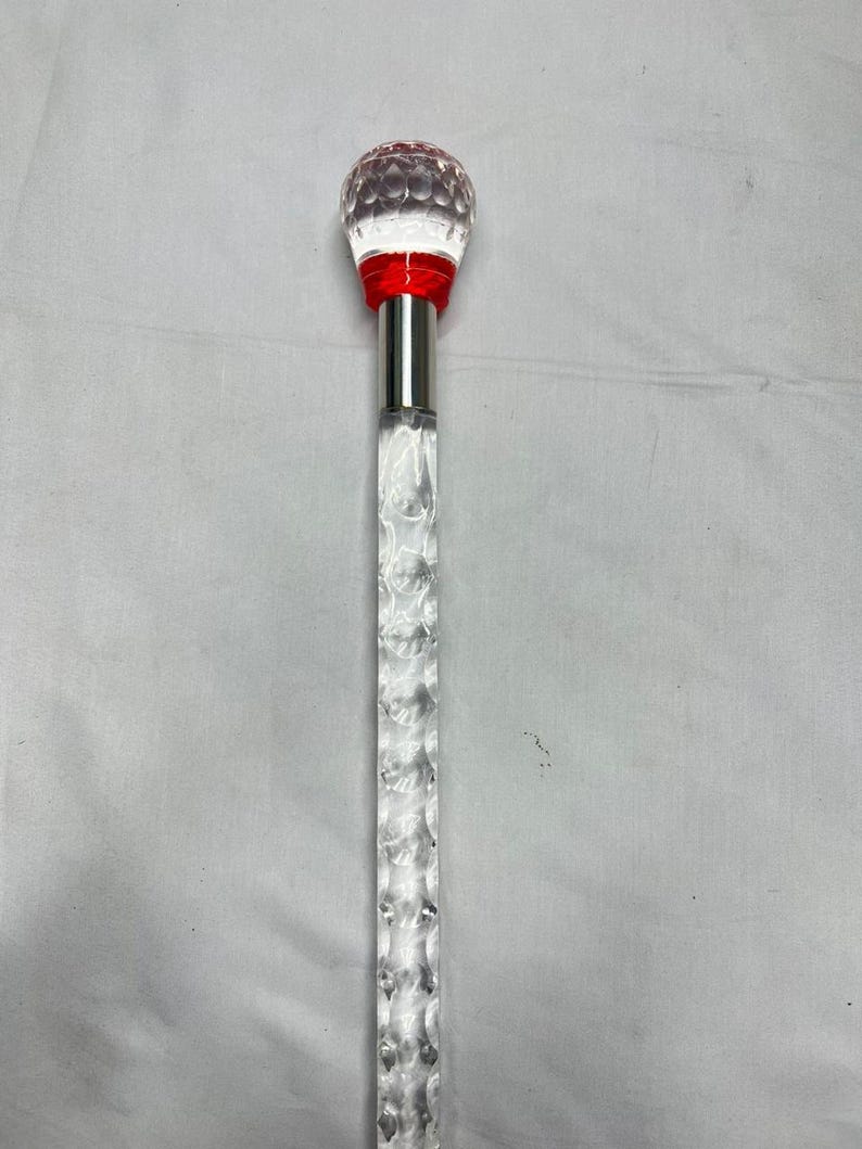 May include: A clear, decorative cane with a faceted, crystal-like shaft and a round, textured top. The top is clear with red accents and a silver metal collar. The cane is designed for decorative purposes.
