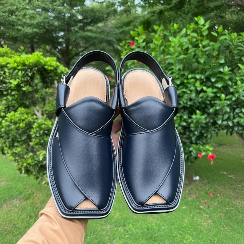 May include: A pair of navy blue leather Peshawari sandals with a white trim. The sandals have a strap around the ankle and a square toe design. The background is a blurred view of green foliage.
