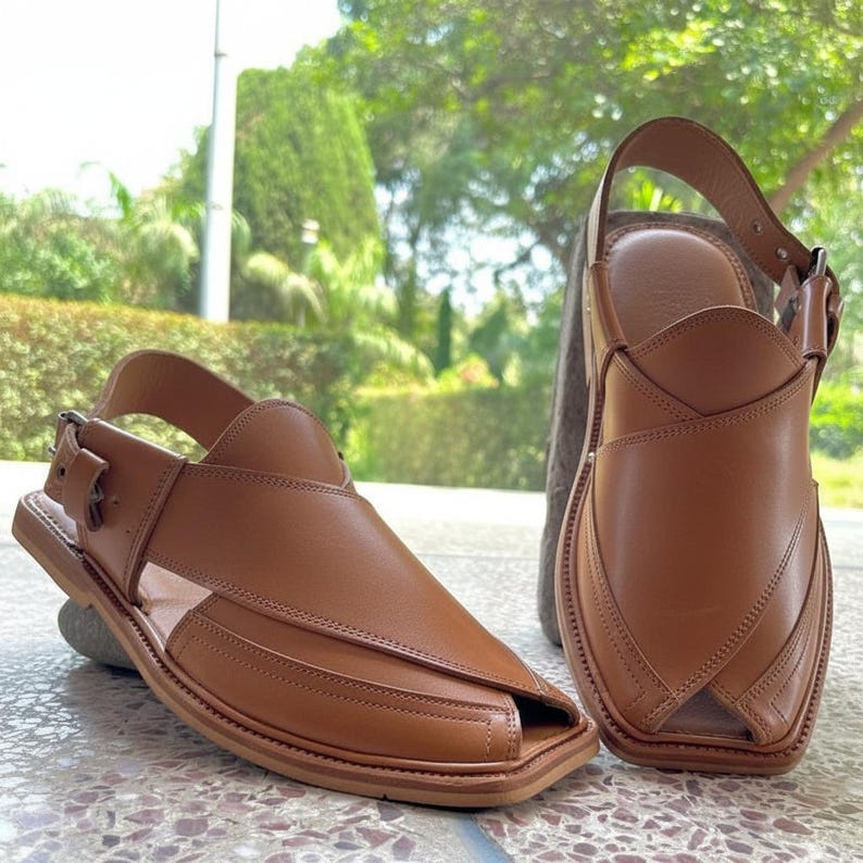 May include: A pair of brown leather sandals with a square toe and a strap that buckles around the ankle. The sandals have a simple, classic design and are made of smooth, high-quality leather. The sandals are displayed on a stone surface.