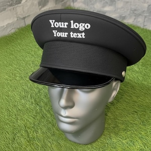 May include: Black hat with a shiny black visor, displayed on a mannequin head. The hat features the text "Your logo Your text" in white. The hat has a textured band and a small silver button on the side. The background is green and gray.