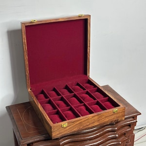 May include: A wooden watch box with a hinged lid, featuring a rich, reddish-brown exterior and a deep red velvet interior. The box is divided into multiple compartments, each designed to hold a watch. Gold-colored clasps secure the lid.