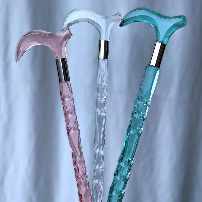 May include: Three decorative walking canes in pink, clear, and teal. Each cane features a curved handle and a textured shaft with a silver band. The canes are designed for style and support.