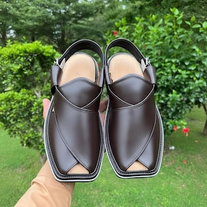 May include: A pair of brown leather Peshawari sandals with a white trim and a strap. The sandals have a square toe and a leather insole. The background is a green, leafy environment.
