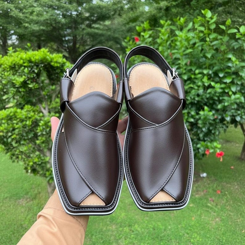 May include: A pair of brown leather Peshawari sandals with a white trim and a strap. The sandals have a square toe and a leather insole. The background is a green, leafy environment.