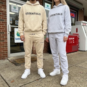 May include: A beige hooded sweatshirt and matching sweatpants set with black ESSENTIALS text on the chest and leg, alongside a light grey hooded sweatshirt and matching sweatpants set with black ESSENTIALS text on the chest and leg.