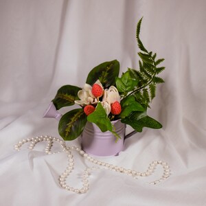 May include: A small, lavender watering can filled with white and red flowers and green foliage. A strand of white pearls is draped in front of the watering can. The background is a white fabric.