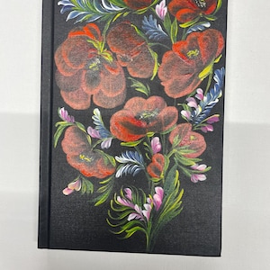 Op de afbeelding: Een zwart notitieboek met een levendig bloemmotief. De omslag toont een boeket van rode klaprozen, groene bladeren en accenten van blauw en roze. Het kunstwerk is geschilderd in een volkskunststijl.