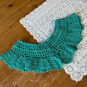 May include: Two pieces of turquoise crochet lace, possibly a collar or trim, are displayed on a wooden surface next to a white, eyelet-embroidered cloth. The crochet work features an open, geometric design with a scalloped edge.