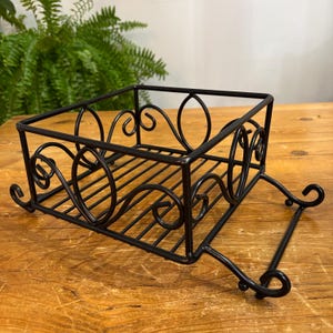May include: A black metal decorative basket with a rectangular shape and ornate scrollwork design. The basket has a slatted base and a handle-like structure on the bottom. The basket is sitting on a wooden surface.