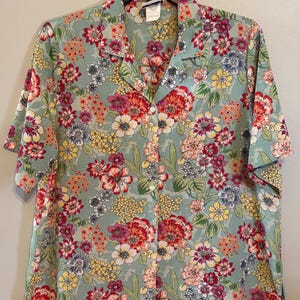 May include: A short-sleeved button-down shirt with a floral pattern. The shirt features a light green background with a vibrant mix of red, pink, yellow, and blue flowers. The shirt is hanging on a black hanger.