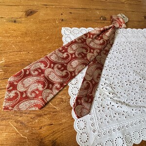 May include: A rust-colored paisley tie with a cream-colored pattern, lying on a white lace doily. The tie is made of a textured fabric and is knotted at the top. The background is a wooden surface.