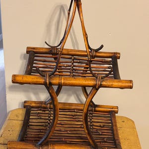 May include: A vintage bamboo magazine rack with a handle. The rack has two tiers and is constructed with woven bamboo slats and curved supports. The bamboo has a warm, natural color with darker accents.