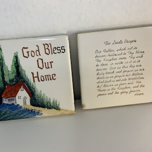 May include: Two white ceramic tiles with a painted image of a house and trees on one tile and the Lord's Prayer on the other. The tile with the house has the words "God Bless Our Home" written on it.