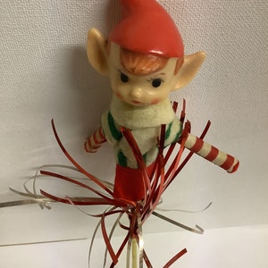 May include: A vintage Christmas elf ornament with a red hat, green and white striped shirt, and red pants. The elf is holding a candy cane and is attached to a gold and red wire streamer.