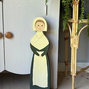 May include: A wooden figurine of a woman wearing a white bonnet, a green dress, and a white apron. The figurine is painted with a smiling face and has a simple, folk art style.