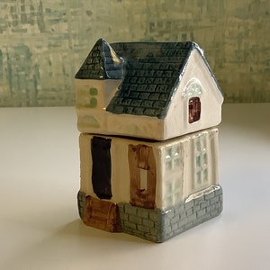 May include: A small ceramic house shaped trinket box with a blue roof, white walls, and brown accents. The house has a small window and a door with a brown step.