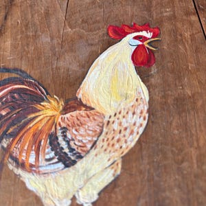 May include: A hand-painted rooster on a wooden surface. The rooster is yellow with brown and white markings. It has a red comb and wattle and is facing to the right.
