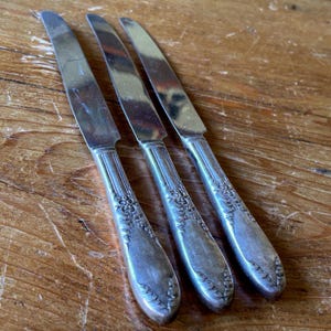 May include: Three silver knives with ornate handles are arranged diagonally on a wooden surface. The knives have a classic design with a decorative pattern on the handles and a shiny, reflective blade.